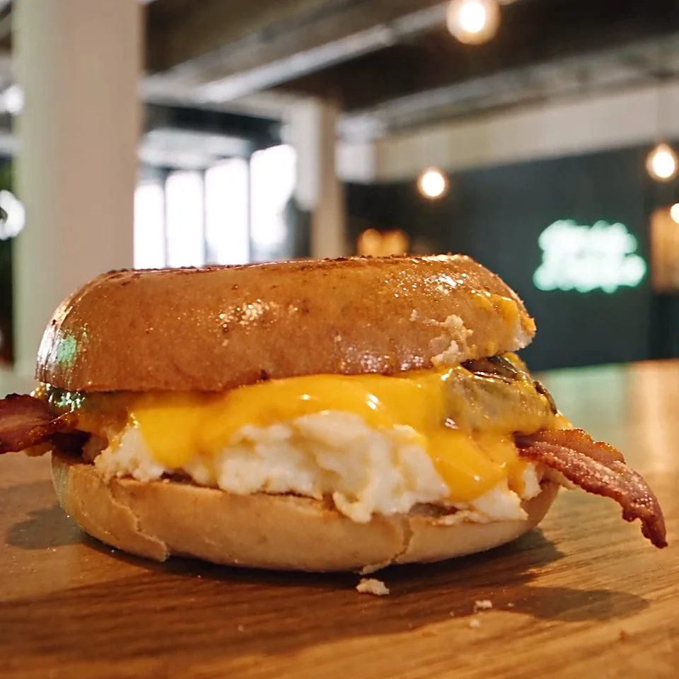 Breakfast Bagel mit Spiegelei, Bacon und geschmolzenem Cheddar auf dem Holztisch im Café